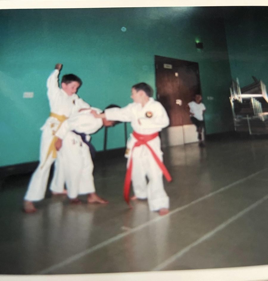 Two young boys in white karate suits practising together in a sports hall. Ryan, on the left, wears a yellow belt; James, on the right, wears a red belt.